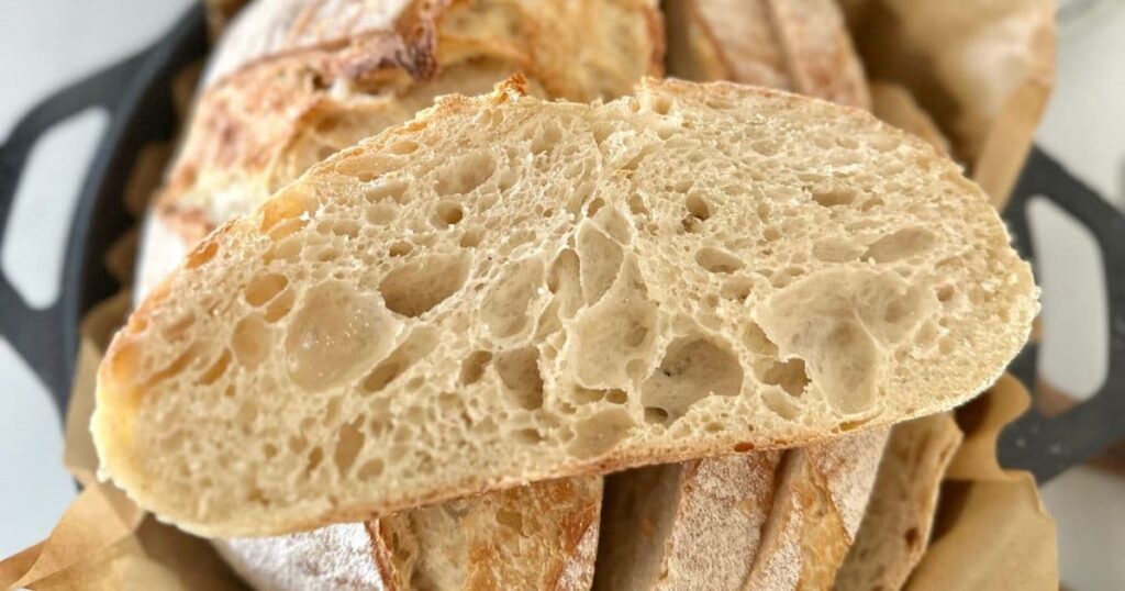 a slice of sourdough bread made with all purpose flour photographed on top of a loaf which has been sliced. You can see a black Lodge Dutch oven underneath the loaf.