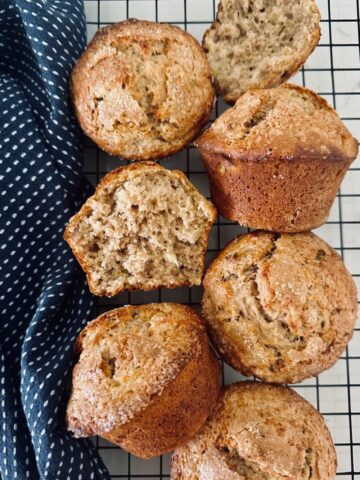 6 sourdough banana muffins sitting on a black cooling rack. There is a blue dish towel on the left of the muffins.