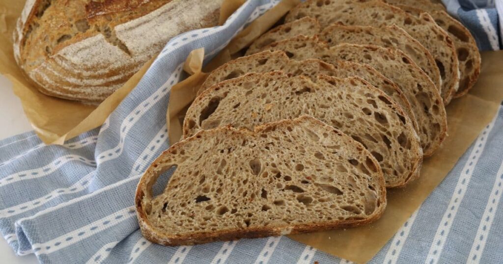 Sourdough country loaf that has been sliced up and laid on a blue striped dish towel. There is also a whole sourdough country loaf in the background of the photo.