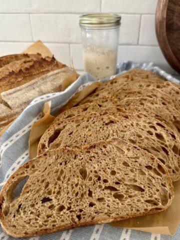 Loaf of sourdough country bread that has been sliced and laid on a blue striped dish towel. There is a jar of sourdough starter in the background.