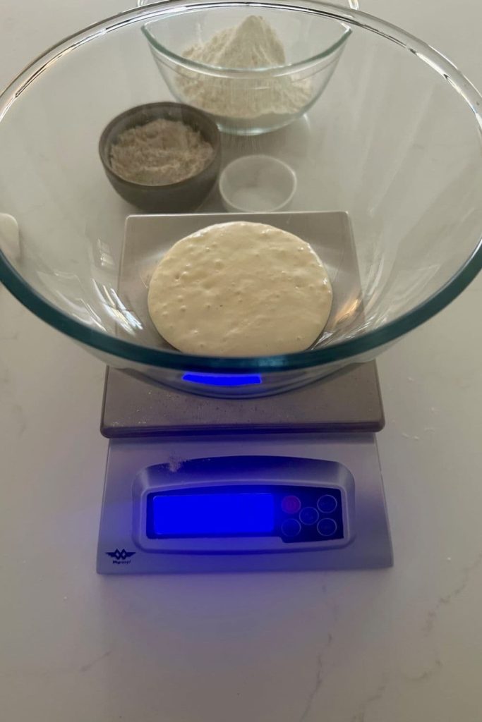 A glass mixing bowl sitting on a digital baking scale. There is 100g of sourdough starter in the bowl and you can see a bowl of bread flour, whole wheat flour and salt in the background.