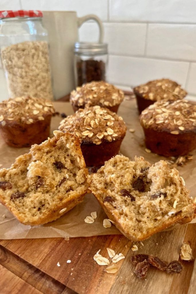 A close up of a sourdough oatmeal raisin muffin that has been broken open to show the inside. There are more muffins in the background, along with some rolled oats and raisins in glass jars.
