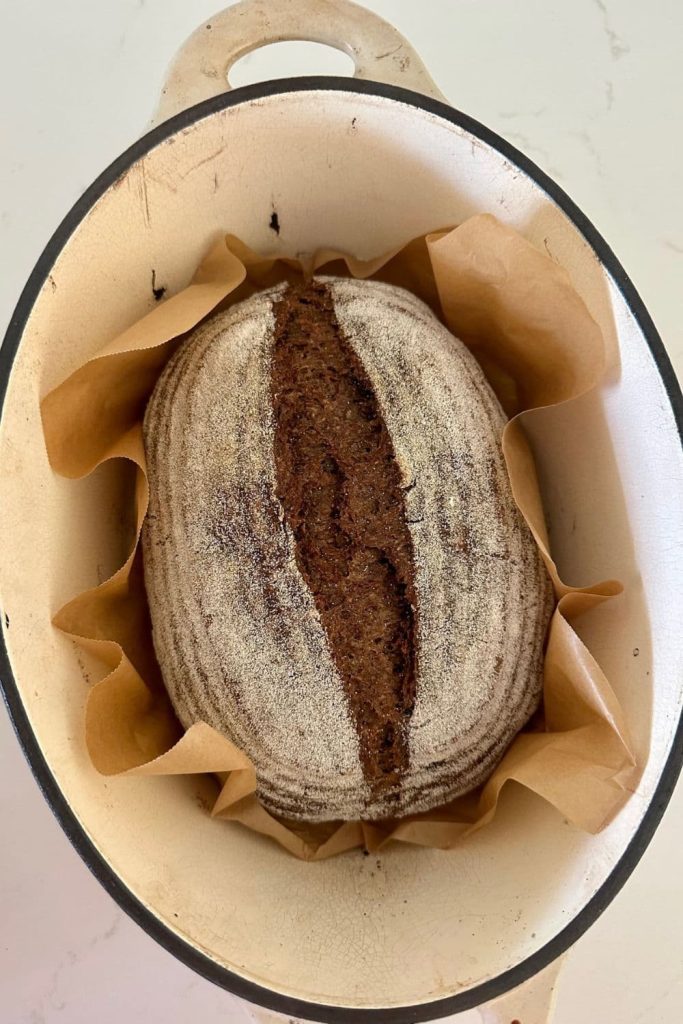Loaf of sourdough pumpernickel bread nestled in a piece of parchment paper inside a cream enamel Dutch Oven.