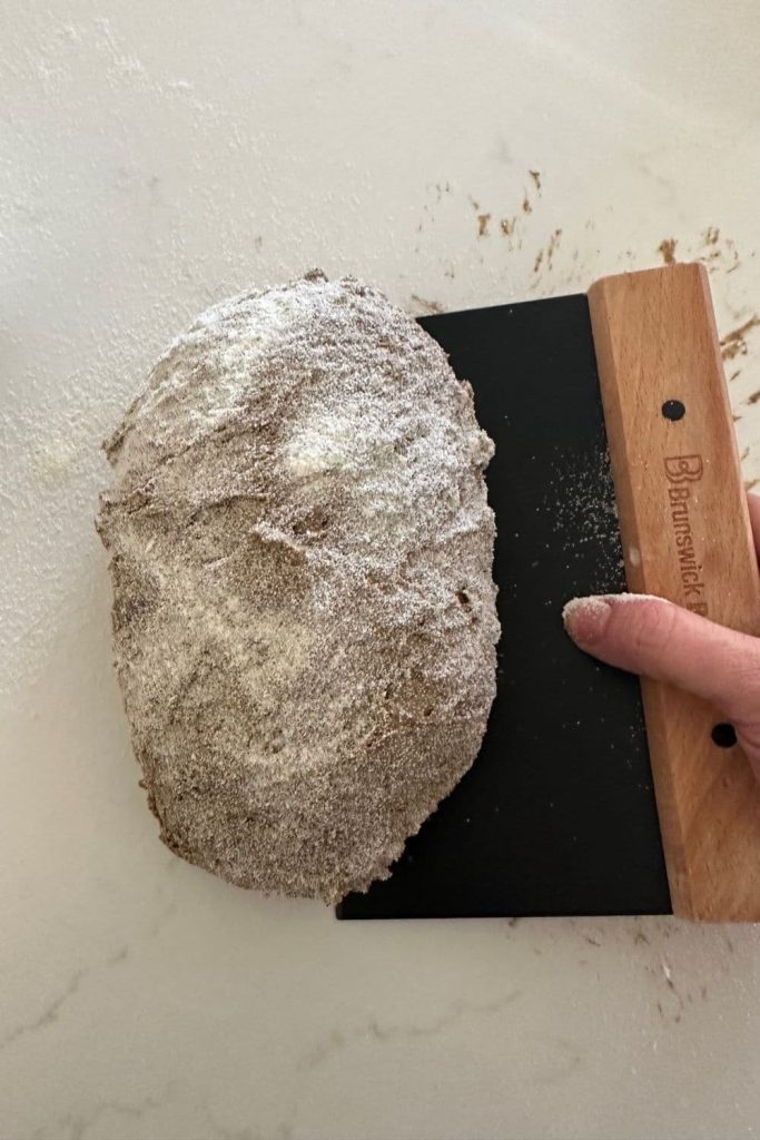 sourdough pumpernickel dough being lifted up with a black and wooden handled bench scraper. The dough has been dusted in semolina flour.