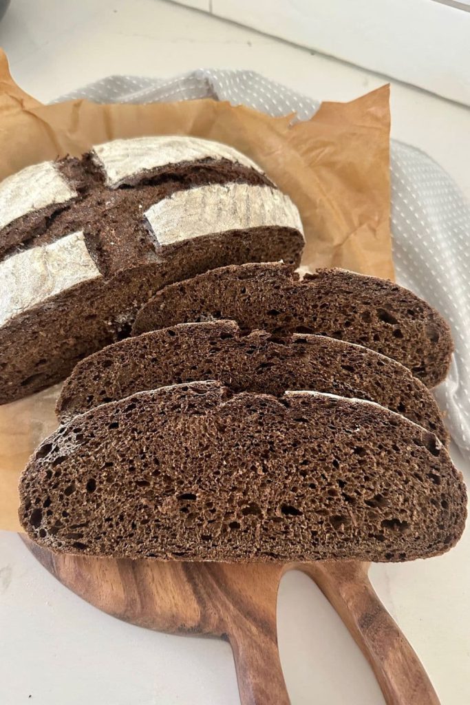 3 slices of sourdough pumpernickel bread that have been laid on a wooden board. The rest of the pumpernickel boule can be seen sitting in the background.
