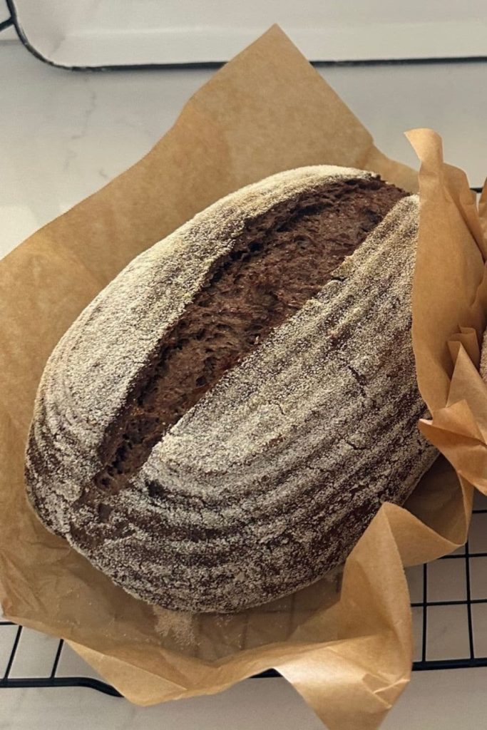 Loaf of sourdough pumpernickel nestled in a piece of parchment paper. It has just come out of the oven and is sitting on a wire cooling rack.