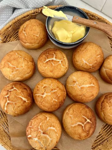 A large cane basket containing 11 sourdough tiger rolls all laid out so you can see the crackly crust. There is a dish of butter and a small butter spreader in the basket also.