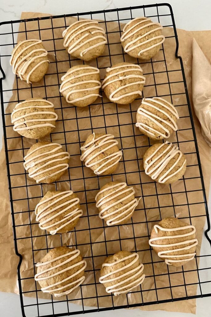 15 soft sourdough discard pumpkin spice cookies on a black wire cooling rack. There is a piece of parchment paper underneath the rack and a piping bag of glaze to the right.