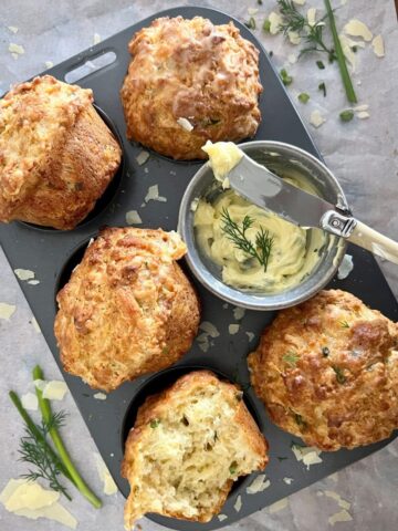 Sourdough Cheese Muffins sitting in a muffin tin. There is a bowl of herbed butter sitting in the muffin tin. There is dill and parmesan cheese sprinkled around the muffins.