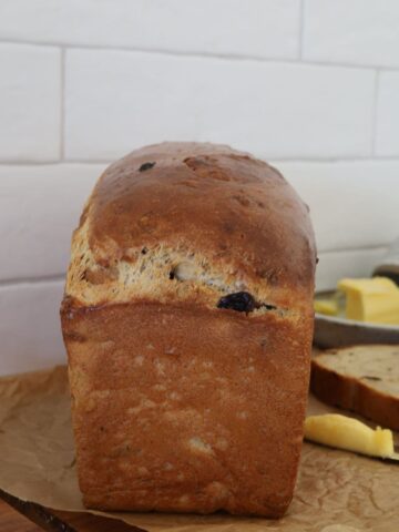 Whole loaf of sourdough discard cinnamon raisin bread sitting on a wooden board. There is a butter dish and butter knife in the background of the photo.