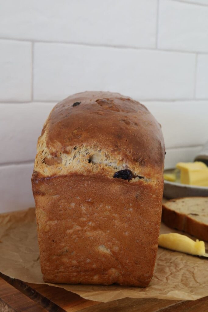 Whole loaf of sourdough discard cinnamon raisin bread sitting on a wooden board. There is a butter dish and butter knife in the background of the photo.