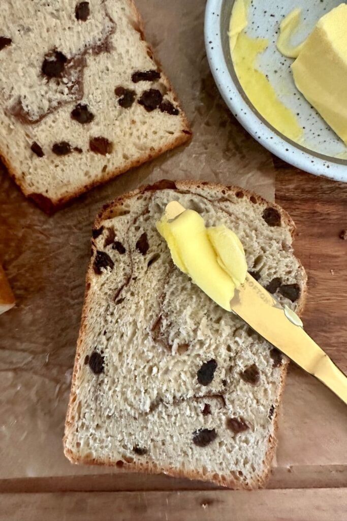 2 slices of sourdough discard cinnamon raisin bread laying on a wooden board. There is a gold knife with butter laying on one of the slices and you can see a butter dish in the top right corner of the photo.