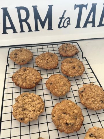 A black wire cooling rack with some sourdough oatmeal raisin cookies cooling. There is a farm to table sign in the background of the photo.