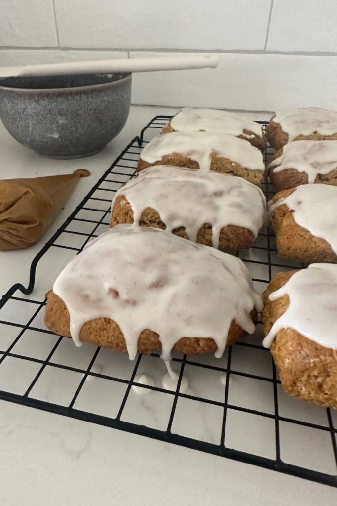 Sourdough pumpkin scones that have been drizzled with vanilla glaze. They are sitting on a black wire rack and there is a grey bowl and spatula in the background.