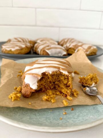 Sourdough pumpkin scone with vanilla glaze and pumpkin spice glaze sitting on a blue stoneware plate. There are a whole plateful of sourdough pumpkin scones in the background.