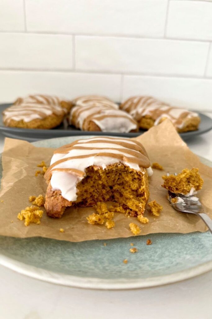 Sourdough pumpkin scone with vanilla glaze and pumpkin spice glaze sitting on a blue stoneware plate. There are a whole plateful of sourdough pumpkin scones in the background.