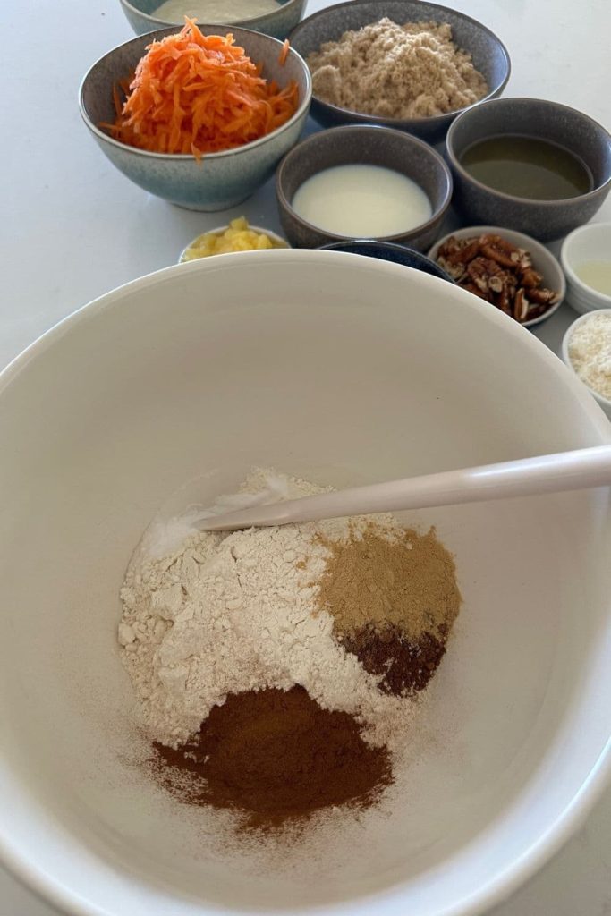 Dry ingredients used to make sourdough carrot cake in a large cream mixing bowl. You can see the remaining ingredients in the background of the photo.