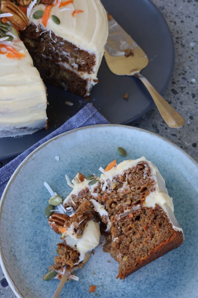 Slice of sourdough carrot cake on a blue stoneware plate. You can see the rest of the sourdough carrot cake in the background of the photo.
