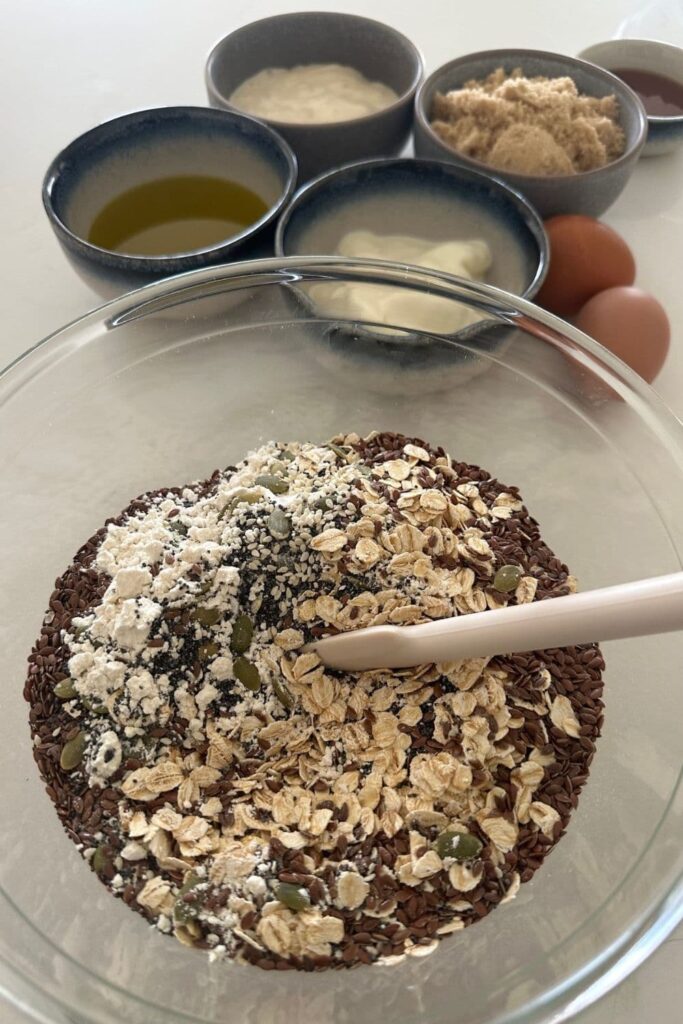 Dry ingredients for seedy sourdough breakfast loaf in a glass mixing bowl with a white jar spatula.