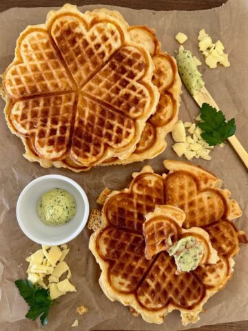 Flat lay of sourdough cheese waffles sitting on a wooden board. There is a dish of garlic butter sitting next to the waffles and the display is garnished with parsley.