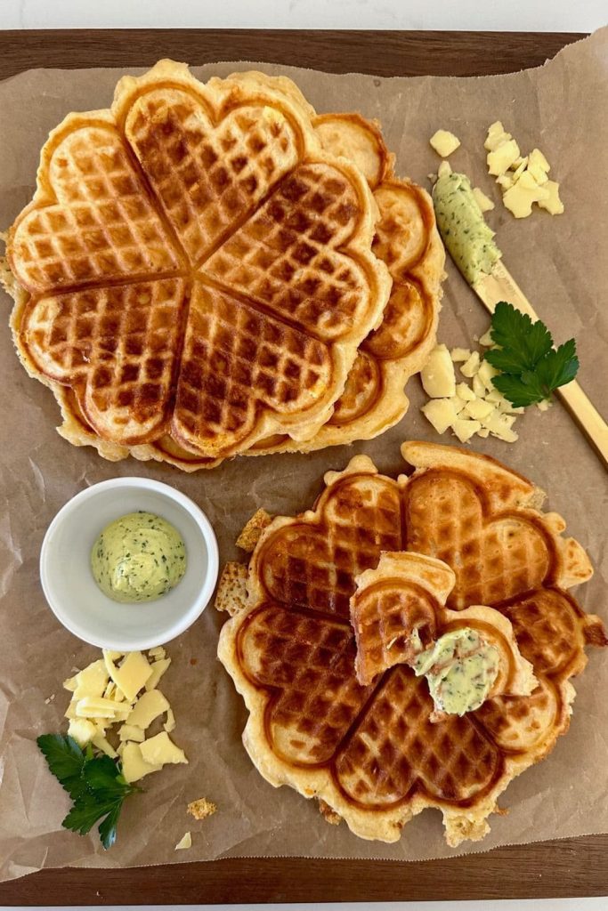 Flat lay of sourdough cheese waffles sitting on a wooden board. There is a dish of garlic butter sitting next to the waffles and the display is garnished with parsley.