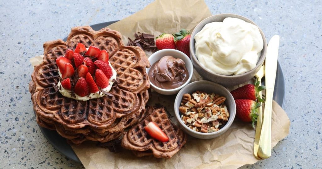 Sourdough chocolate waffles stacked up and topped with whipped cream and fresh strawberries. There are bowls of nutella, pecans, fresh strawberries and whipped cream placed onto a platter around the sourdough chocolate waffles.