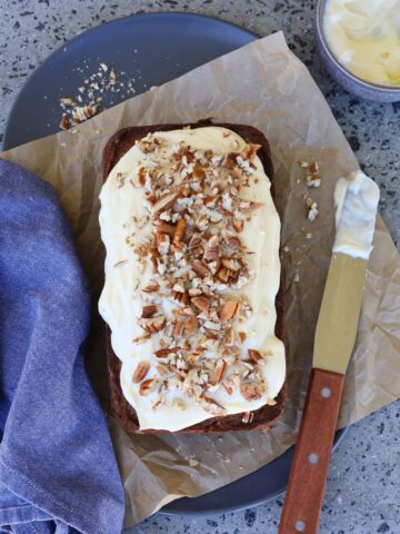 sourdough gingerbread loaf cake photographed from above. It is iced with vanilla cream cheese frosting and has a palette knife sitting to the right and a blue dish towel sitting to the left.