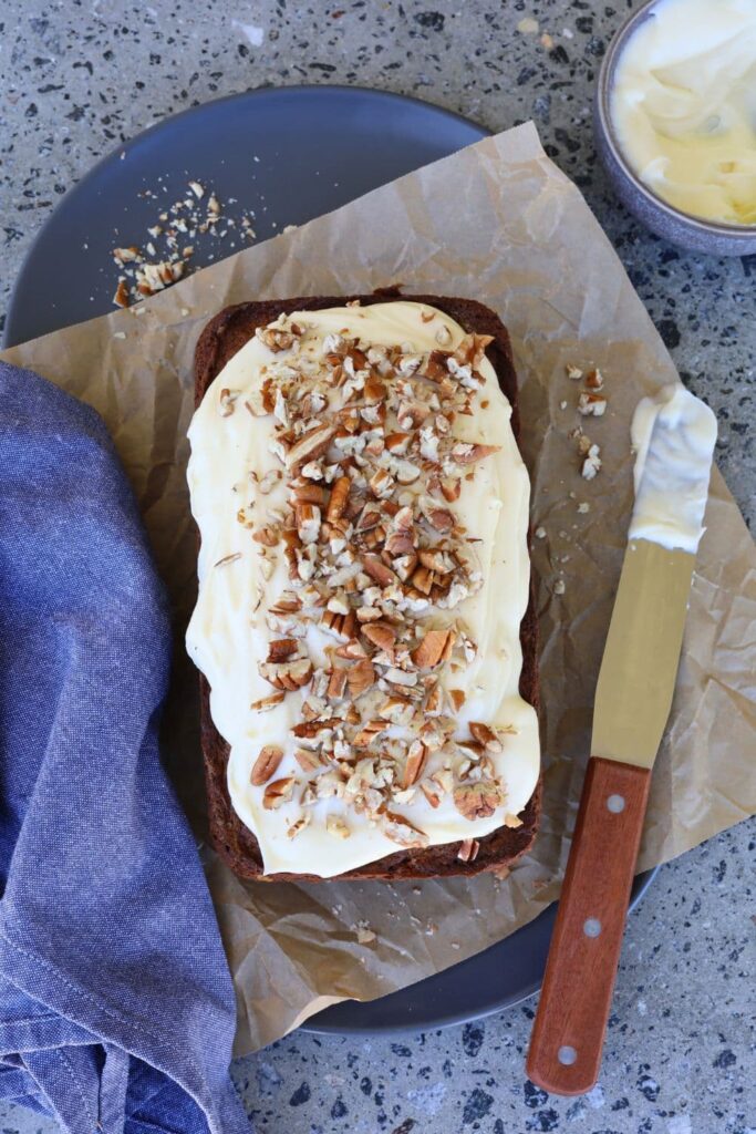 sourdough gingerbread loaf cake photographed from above. It is iced with vanilla cream cheese frosting and has a palette knife sitting to the right and a blue dish towel sitting to the left.