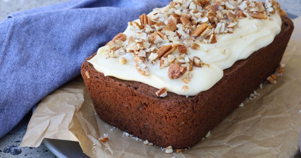 Sourdough gingerbread loaf cake sitting on a piece of parchment paper. It is frosted with vanilla cream cheese icing and sprinkled with pecan nuts.