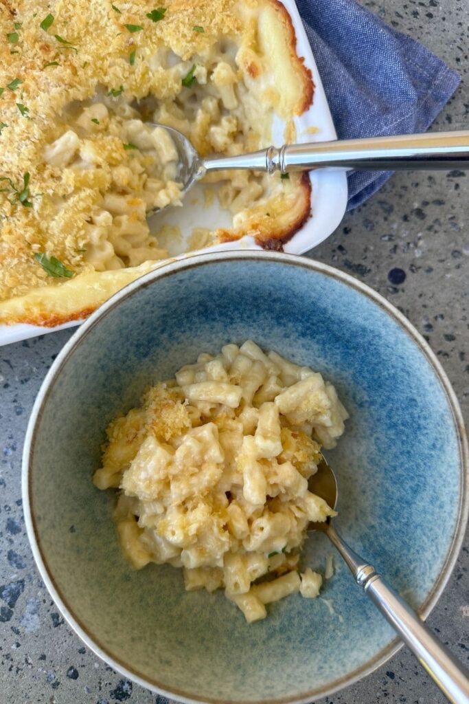 Sourdough mac and cheese served in a blue stoneware bowl. You can see the rest of the mac and cheese in a white baking dish behind the individual serving.