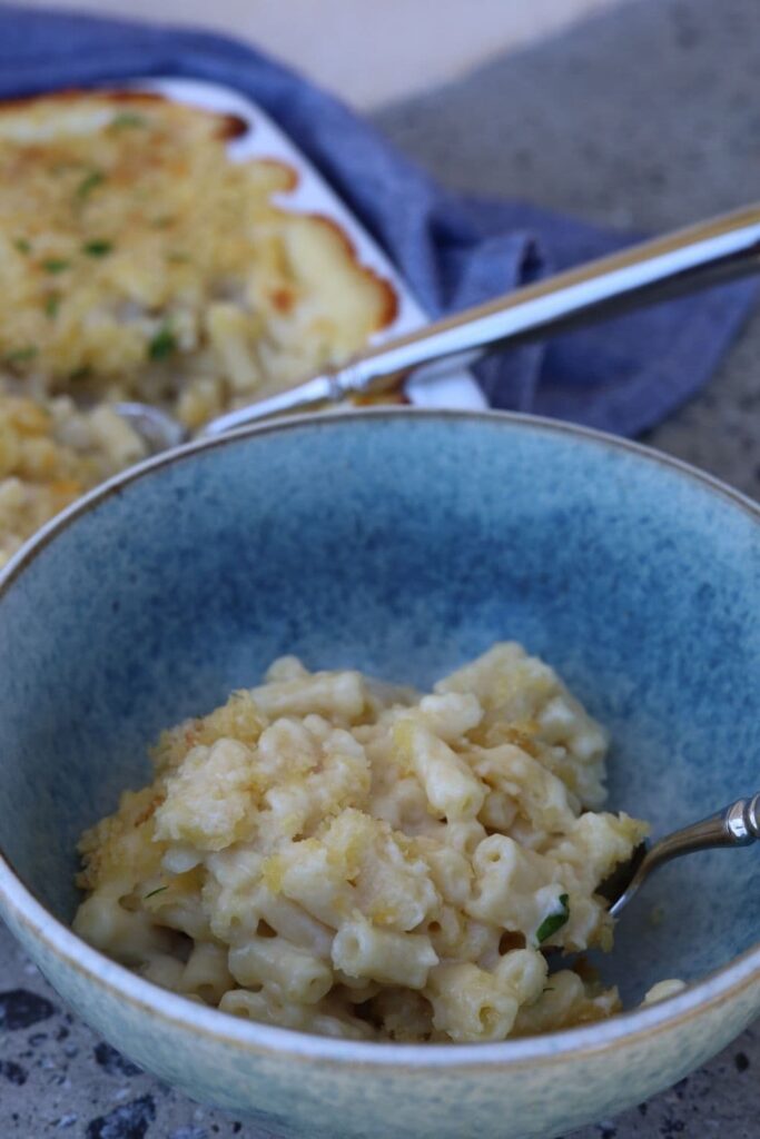 Sourdough mac and cheese served in a blue stoneware bowl. You can see the rest of the mac and cheese in a white baking dish behind the individual serving.