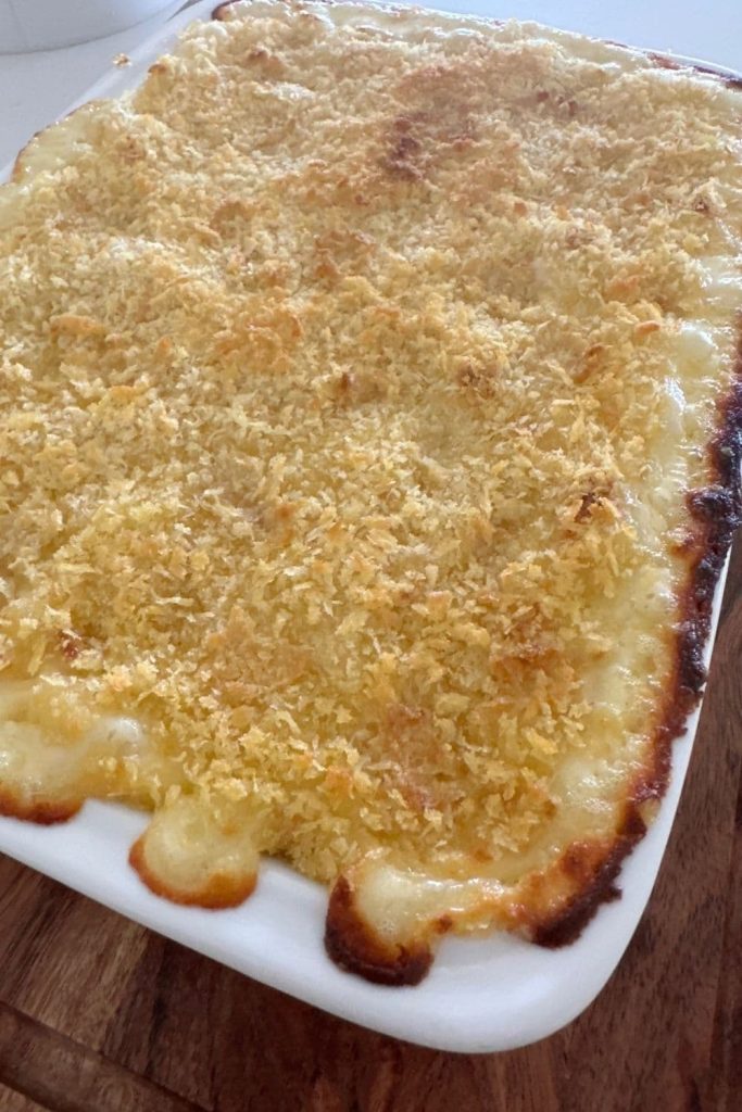 Sourdough mac and cheese baked in a white oven dish with a crumb topping. The molten cheese sauce has oozed out the sides and over browned.