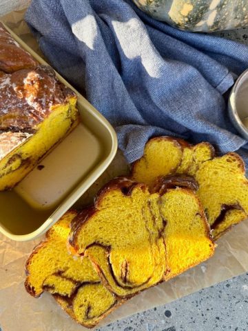 Sourdough pumpkin babka that has been sliced and laid out on a piece of parchment paper. You can see the remainder of the loaf sitting in a gold loaf pan. There is a blue dish towel and a whole pumpkin in the left hand side of the photo.
