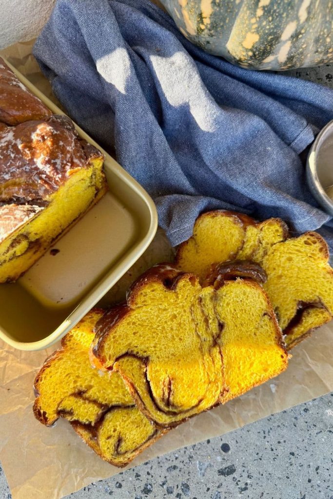 Sourdough pumpkin babka that has been sliced and laid out on a piece of parchment paper. You can see the remainder of the loaf sitting in a gold loaf pan. There is a blue dish towel and a whole pumpkin in the left hand side of the photo.