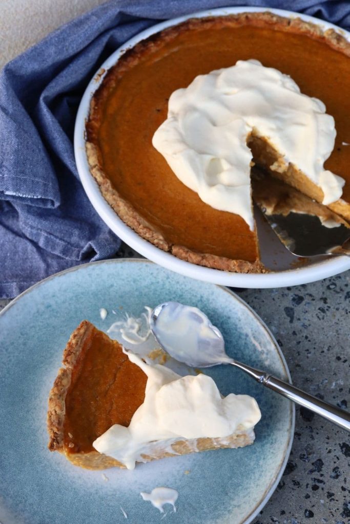A slice of sourdough pumpkin pie served on a blue stoneware plate topped with whipped cream. You can see the rest of the sourdough pumpkin pie in the background of the photo.