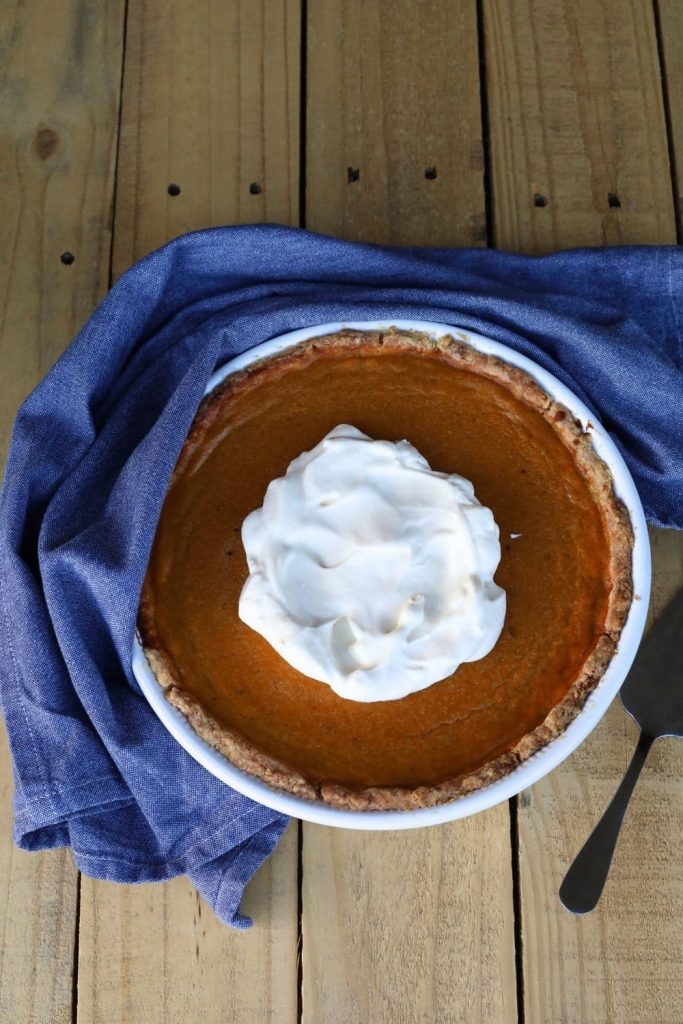 Sourdough pumpkin pie topped with whipped cream and baked in a white pie dish. It is sitting on a wooden farmhouse table with a blue dish towel surrounding the pie plate.