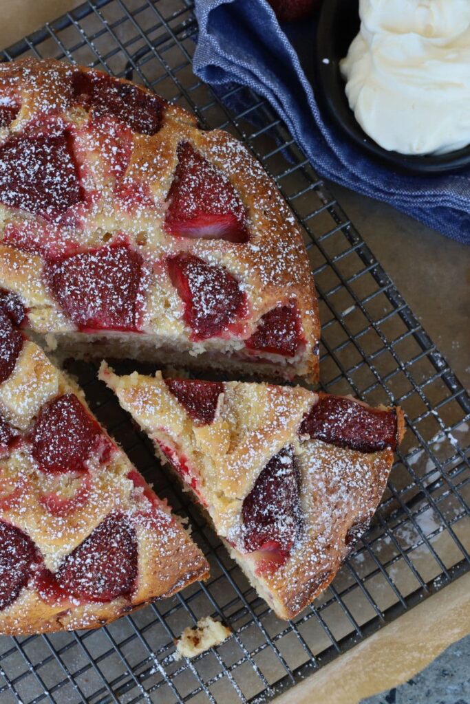 A round sourdough strawberry cake sitting on a wire cake rack. The cake has been dusted with icing sugar and a slice has been cut out of the cake.