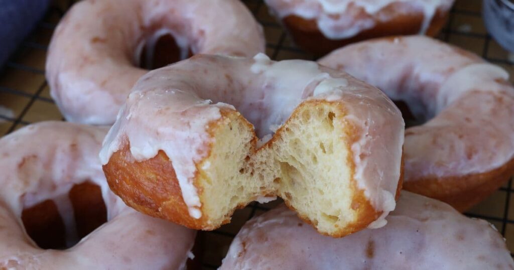 Fried sourdough donuts coated in vanilla glaze. There is a bite taken out of the donut so you can see the light, airy interior.