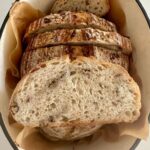 A loaf of high protein sourdough bread that has been sliced up and put back inside the Dutch Oven. There is a slice sitting on top of the loaf so you can see the gorgeous lacy, airy crumb studded with nuts and seeds.