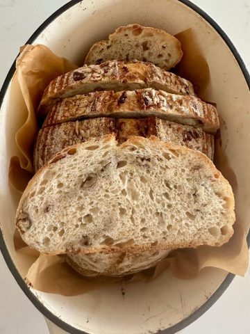 A loaf of high protein sourdough bread that has been sliced up and put back inside the Dutch Oven. There is a slice sitting on top of the loaf so you can see the gorgeous lacy, airy crumb studded with nuts and seeds.