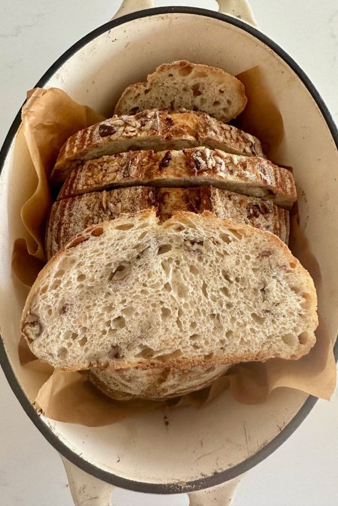 A loaf of high protein sourdough bread that has been sliced up and put back inside the Dutch Oven. There is a slice sitting on top of the loaf so you can see the gorgeous lacy, airy crumb studded with nuts and seeds.