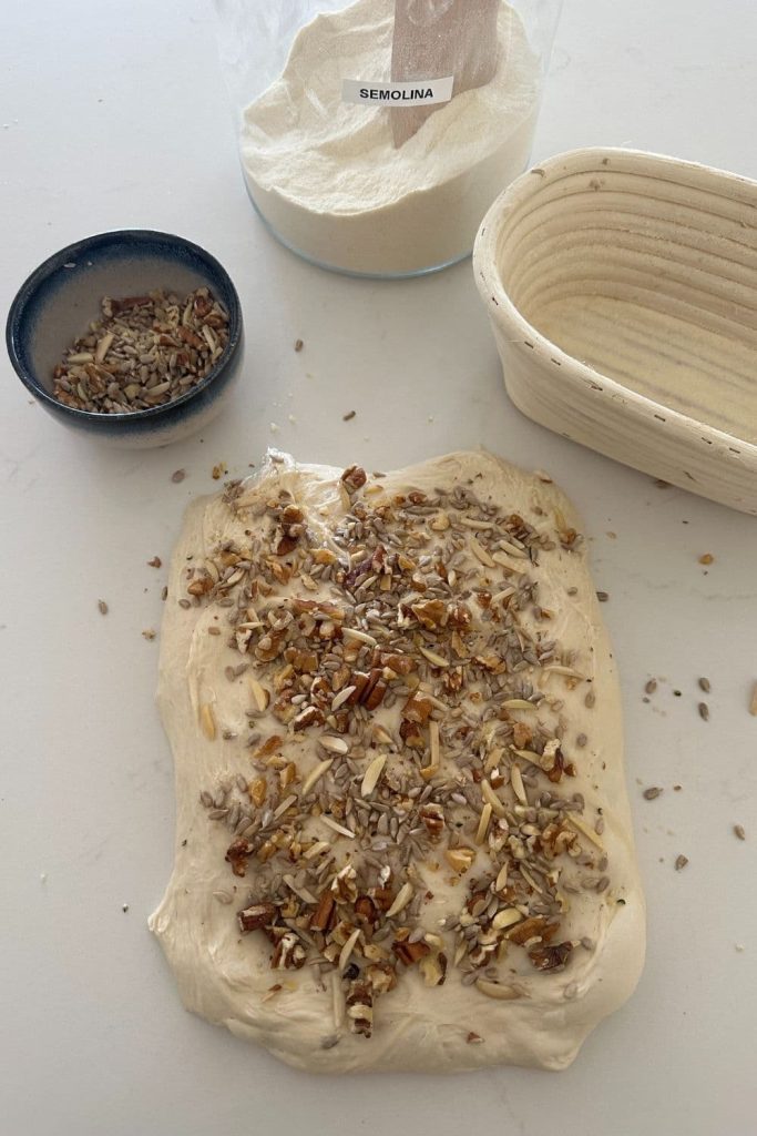 Dough for a high protein sourdough bread loaf laid out in a rectangle and covered with a high protein blend of nuts and seeds. There is a also a banneton and jar of semolina.