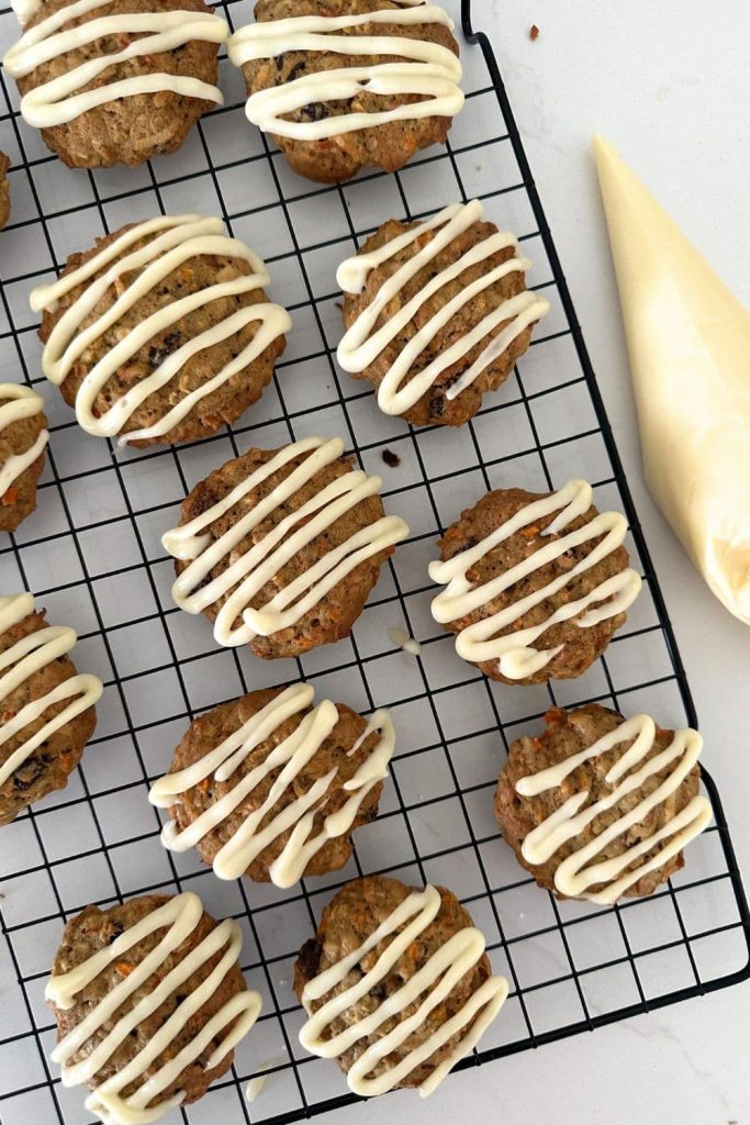 Sourdough carrot cake cookies sitting on a wire cooling rack. They have been drizzled with vanilla cream cheese glaze using a piping bag.