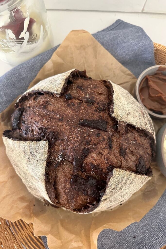 Round loaf of sourdough chocolate bread sitting on a piece of parchment paper and a blue dish towel.