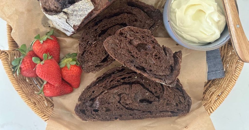Loaf of sourdough chocolate bread that has been sliced and displayed in front of the whole loaf. There is caramel, whipped cream and strawberries in the flat lay display.