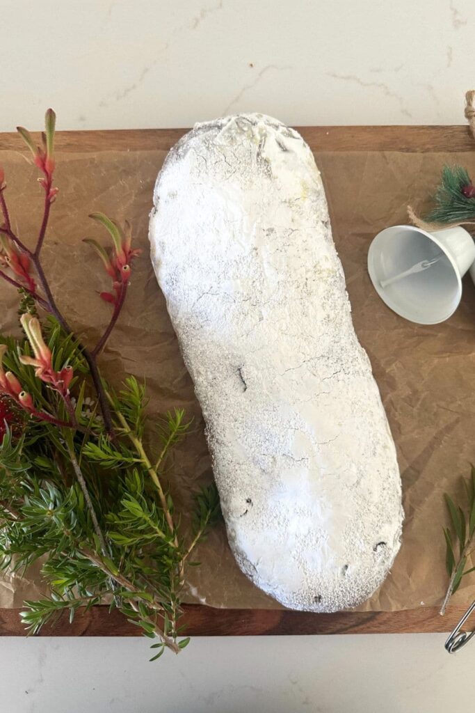 A traditional sourdough stollen dusted in white powedered sugar. There is a bunch of red and white Christmas flowers to the left of the sourdough stollen.