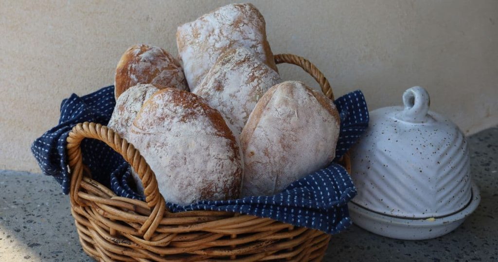 A basket of sourdough cibatta rolls that are heavily floured on the outside. The basket is lined with a dark blue dish towel and there is a cream stoneware butter dish sitting to the right of the basket.