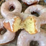 Fried sourdough donuts with vanilla glaze laid out on a wooden board. One of the donuts has a bite taken out so you can see the light, yeasty inside of the donut.