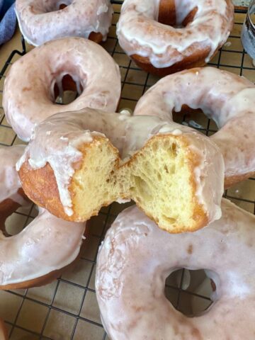 Fried sourdough donuts with vanilla glaze laid out on a wooden board. One of the donuts has a bite taken out so you can see the light, yeasty inside of the donut.