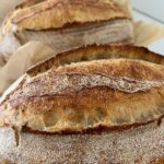 3 loaves of crusty sourdough bread sitting in a row. Each loaf has some parchment paper around it.